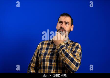 Frognant homme pensant exprimant des doutes et des préoccupations. Petit garçon avec barbe en chemise à carreaux bleu isolée sur fond bleu studio Banque D'Images