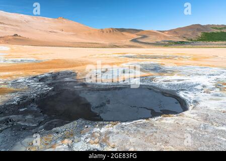 Piscine de boue bouillonnante dans une zone géothermique en Islande, par une belle journée d'été. Une route sinueuse grimpant une colline volcanique aride est visible en arrière-plan. Banque D'Images