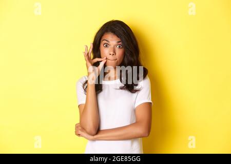 Portrait d'une jeune femme afro-américaine qui promet de garder le secret, de sceller les lèvres, de zipper la bouche avec les doigts, debout sur fond jaune Banque D'Images