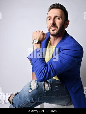 Portrait de l'homme moderne adulte non rasé dans un vêtement moderne et décontracté, assis sur le côté du tabouret avec les jambes croisées Banque D'Images