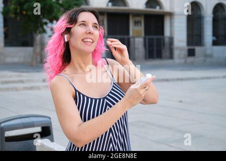 Jeune femme caucasienne aux cheveux roses mettant des écouteurs sans fil dans la rue. Banque D'Images