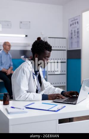 Portrait d'un médecin afro-américain assis au bureau à l'aide d'un ordinateur portable pour examen dans un cabinet médical à la clinique de santé. Black man avec périphérique moderne et fichiers de document sur table Banque D'Images
