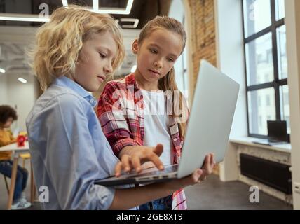 Deux enfants curieux, un petit garçon et une fille utilisant un ordinateur portable tout en se tenant dans une salle de classe pendant la leçon DE STEM Banque D'Images