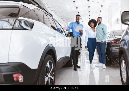 Un jeune couple noir heureux achète une nouvelle voiture ensemble, en parlant avec un vendeur sympathique chez un concessionnaire automobile Banque D'Images