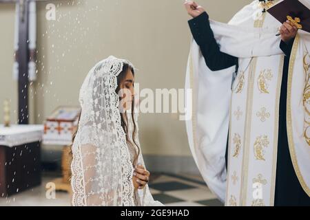 Prêtre de bénédiction avec l'eau sainte mariée élégante dans le mouchoir à l'autel pendant le mariage Saint dans l'église. Cérémonie de mariage dans la cathédrale. Classique spirituel W Banque D'Images