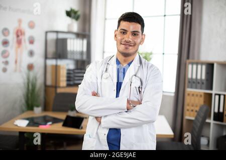 Souriant jeune homme de 30 ans arabe médecin asiatique posant avec les armes croisées dans le bureau. Médecin masculin en costume bleu et manteau blanc, avec un stéthoscope autour du cou, posant à la caméra à l'intérieur Banque D'Images