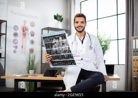 Médecine, radiologie et concept de soins de santé. Un homme souriant, un médecin indien, un manteau blanc, tenant une image IRM et regardant la caméra au-dessus du bureau médical à l'arrière-plan de l'hôpital Banque D'Images