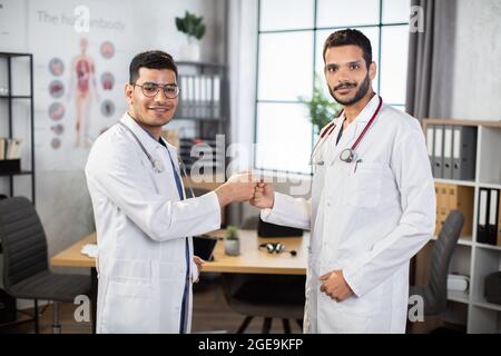 Expérimenté deux médecins indiens asiatiques chirurgiens dans les manteaux médicaux faisant des poings bump, debout dans le bureau moderne de l'hôpital, se préparant à l'opération. Une équipe de deux médecins qui se sont embués de poings Banque D'Images