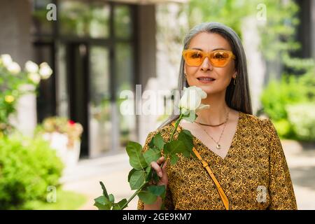 élégante femme d'âge moyen en chemisier à motifs et lunettes de soleil tenant une rose blanche à l'extérieur Banque D'Images