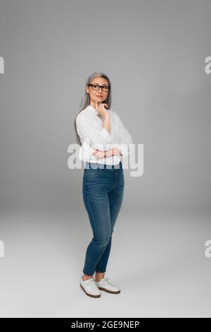 vue sur toute la longueur de la femme mûre en jeans, chemise blanche et lunettes de vue debout sur gris Banque D'Images