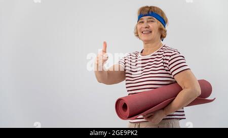 Une femme âgée tient un tapis de yoga et montre son pouce vers le haut. Le retraité approuve les sports. Banque D'Images