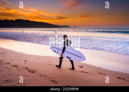 Coucher de soleil sur un surfeur transportant sa planche de surf et marche le long du rivage sur la plage de Fistral à Newquay, en Cornouailles. Banque D'Images