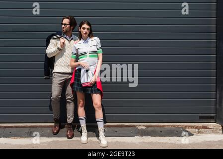 femme en polo à rayures, jupe à carreaux et bottes posant près de l'homme tendance et du mur gris Banque D'Images