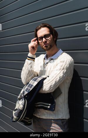 homme tendance en pull blanc ajustement des lunettes près du mur gris à l'extérieur Banque D'Images