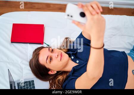 Par-dessus une jeune étudiante positive couché sur le lit et prenant le selfie sur un smartphone tout en ayant une pause pendant des études en ligne à distance à la maison Banque D'Images