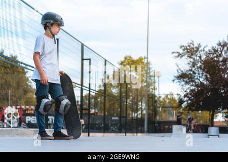 Vue latérale d'un jeune garçon confiant avec planche à roulettes et casque de protection debout dans le parc de skate et regardant loin Banque D'Images