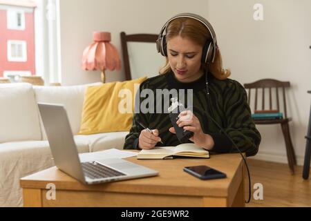 Animateur de radio féminin focalisé avec micro et casque écrivant dans le bloc-notes tout en préparant l'enregistrement de podcast à la maison Banque D'Images