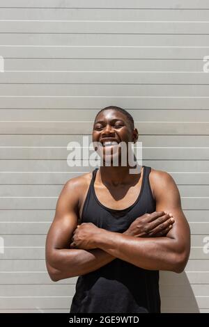 S'adapter à un homme afro-américain debout avec les bras croisés près du mur en métal dans la rue et rire avec les yeux fermés le jour ensoleillé Banque D'Images