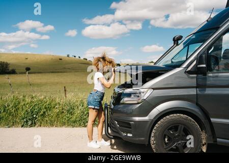 Vue latérale d'une femme en voyage méconnaissable qui regarde sous le capot ouvert d'une camionnette tout en ayant des problèmes pendant le voyage dans la nature estivale Banque D'Images
