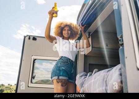 Vue latérale d'une femme noire ravie avec de la bière riant et regardant la caméra tout en se tenant sur l'échelle à l'extérieur de la campeuse et en montant sur le lit le jour d'été Banque D'Images