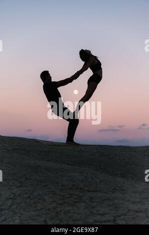 Vue latérale de la silhouette d'une femme flexible méconnaissable debout sur les jambes de l'homme pendant la séance de yoga d'acro sur fond de ciel du soir Banque D'Images