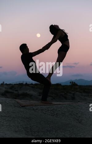 Vue latérale de la silhouette d'une femme flexible méconnaissable debout sur les jambes de l'homme pendant la séance de yoga d'acro sur fond de ciel du soir Banque D'Images