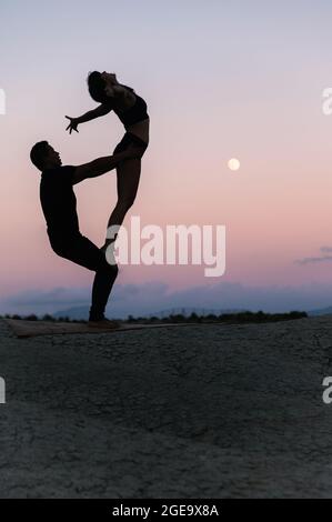 Vue latérale de la silhouette d'une femme flexible méconnaissable debout sur les jambes de l'homme pendant la séance de yoga d'acro sur fond de ciel du soir Banque D'Images