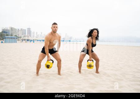Sport multiracial et sportif effectuant un exercice d'oscillation de kettlebell pendant l'entraînement en bord de mer en été Banque D'Images