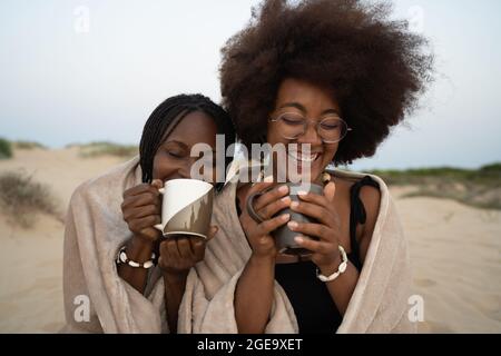 Bonne jeune femme noire meilleure amie avec des tasses de boissons chaudes assis près de la couverture chaude et s'amuser pendant la soirée d'été sur le sable Banque D'Images