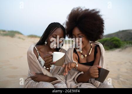 Bonne jeune femme noire meilleure amie avec des tasses de boissons chaudes assis près de la couverture chaude et s'amuser pendant la soirée d'été sur le sable Banque D'Images