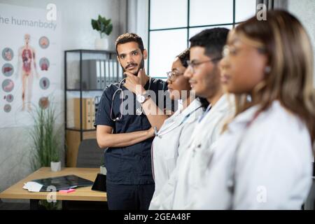 Vue floue du profil de diverses équipes de médecins multiethniques debout dans la salle de clinique moderne. Concentrez-vous sur un spécialiste médical indien arabe compétent et réfléchi, debout derrière vous et regardant l'appareil photo Banque D'Images