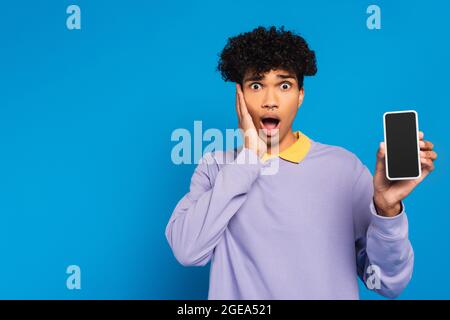 choqué africain américain homme touchant le visage tout en montrant le téléphone portable avec écran blanc isolé sur bleu Banque D'Images