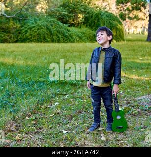 latino garçon debout souriant dans la forêt tenant un ukulele vert à ses côtés. carré Banque D'Images