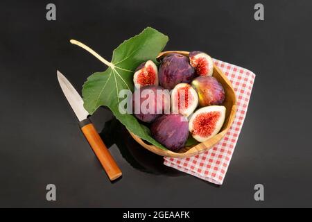 Figues biologiques fraîches et feuille de figues dans un bol en bois avec serviette à carreaux rouges et couteau à découper, sur fond sombre. Figues fraîches. Figues entières et tranchées Banque D'Images