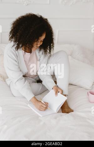 Femme afro-américaine assise sur le lit et prenant des notes dans un journal. Banque D'Images