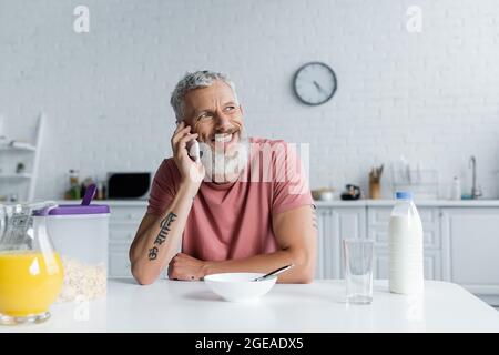 Homme mûr souriant parlant sur un smartphone près du petit déjeuner dans la cuisine Banque D'Images