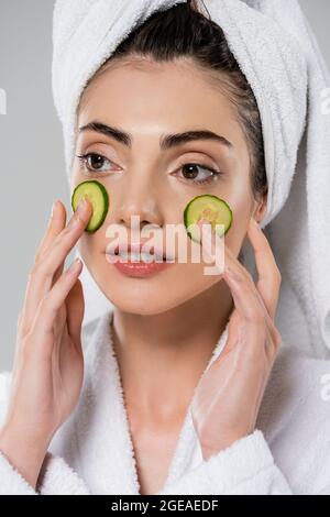 jeune femme avec une serviette sur la tête appliquant le concombre en tranches sur le visage isolé sur le gris Banque D'Images