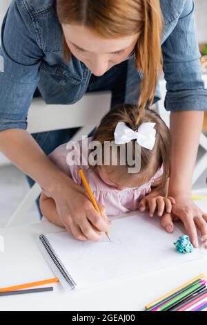 vue en grand angle de l'enseignant aidant une fille handicapée avec le syndrome de down dessin dans la maternelle privée Banque D'Images