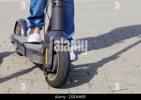 Une fille en Jean fait un scooter électrique dans une rue de la ville, des jambes de femme sur le trottoir. Équitation en été Banque D'Images