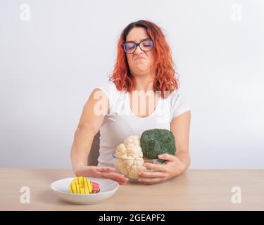 La femme caucasienne préfère une alimentation saine et refuse la restauration rapide. REDHEAD fille choisit entre brocoli et beignes sur fond blanc Banque D'Images