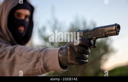 Homme visant avec un pistolet, la nature à l'extérieur arrière-plan, vue rapprochée. Tueur à capuche avec balaclava, tenant un pistolet dans la main gantée, arme de robber concept Banque D'Images