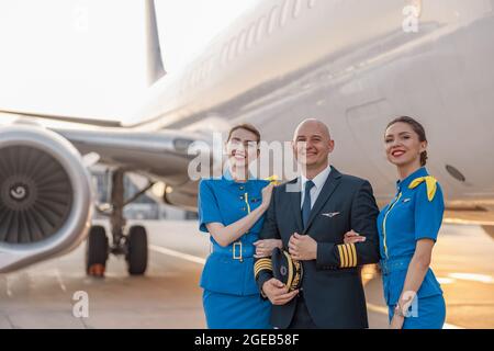 Portrait d'un pilote homme excité posant avec deux hôtesses d'air en uniforme bleu devant un avion à l'aéroport Banque D'Images