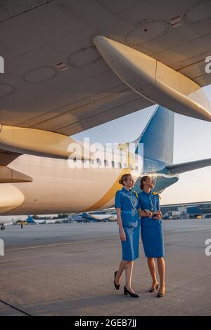 Vue en longueur de deux hôtesses d'air en uniforme bleu souriant, debout devant un gros avion passager à l'aéroport au coucher du soleil Banque D'Images