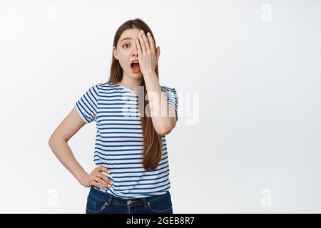 Image de jeune fille sans maquillage couvre la moitié du visage, un côté et regardant surpris à la caméra, debout sur fond blanc Banque D'Images