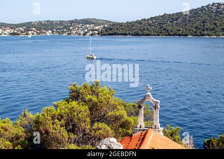 Croix de pierre sur la petite chapelle sur la côte de Rogoznica avec la vue sur la magnifique baie bleue dans le centre de Dalmatie avec des bateaux à voile passant par Banque D'Images