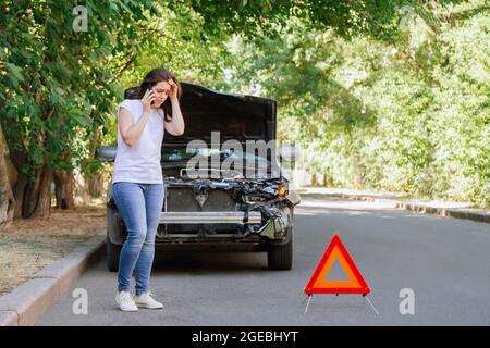 Jeune femme devant une voiture brisée dans un accident de voiture. Femme téléphone appelant pour l'aide et l'assurance après accident de voiture sur la route avec le triangle de circulation Banque D'Images