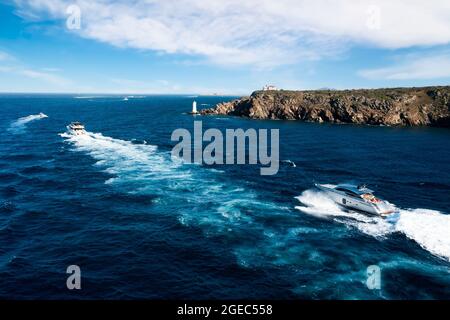 Vue d'en haut, vue aérienne stupéfiante de certains yachts naviguant sur une mer bleue en premier plan et un littoral rocheux avec un phare au loin. Banque D'Images