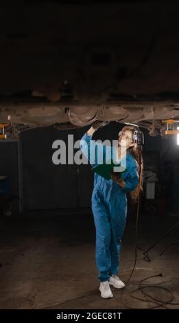 Une belle dame mécanicien de voiture vêtue de combinaisons et de lunettes de sécurité se tient sous la voiture surélevée. Fait une inspection du train de roulement et regarde f Banque D'Images