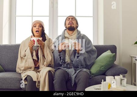 Jeune couple malade enveloppé dans des couvertures assises sur un canapé et éternuant dans des mouchoirs en papier Banque D'Images