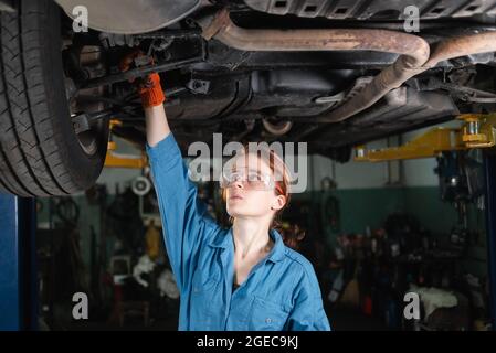 une femme mécanicien de voiture vêtue de lunettes et de combinaisons répare une voiture. Fait tourner le pignon de course avec une clé.4k Banque D'Images